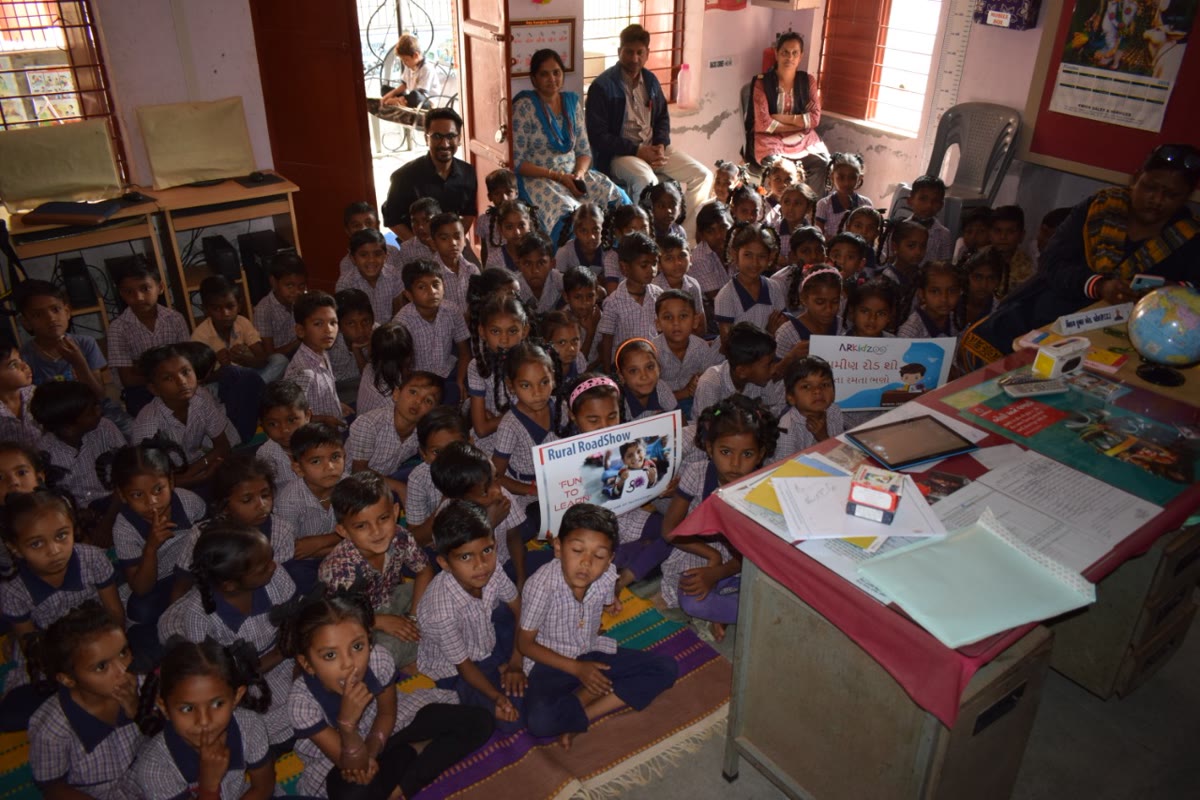 A village classroom full of children beside a Fun To Learn road-show placard